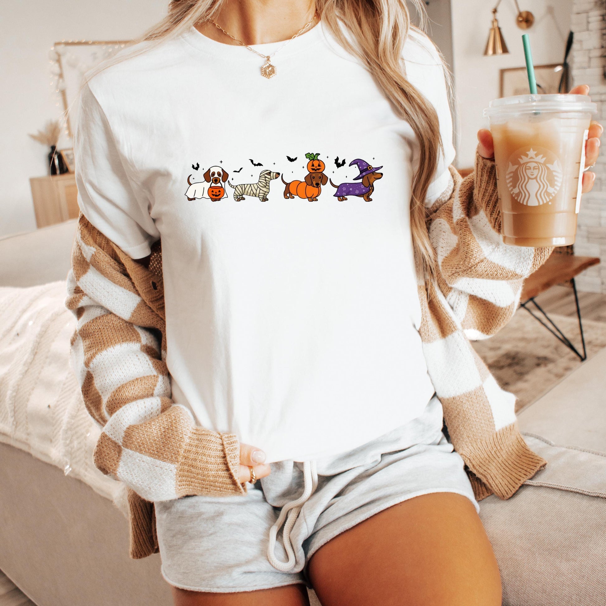 A woman wearing a white t-shirt with a graphic of cartoon dogs and Halloween-themed illustrations, holding a Starbucks drink in her hand.