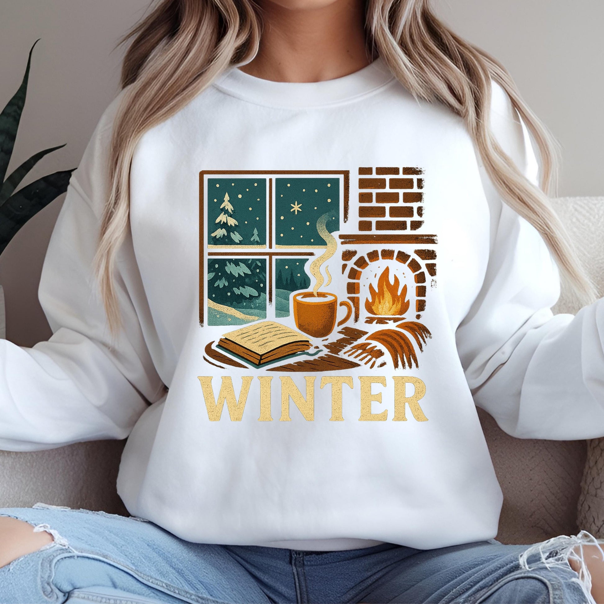 A woman wearing a white sweatshirt with a graphic design depicting a cozy winter scene, including a fireplace, a book, and a cup of coffee.