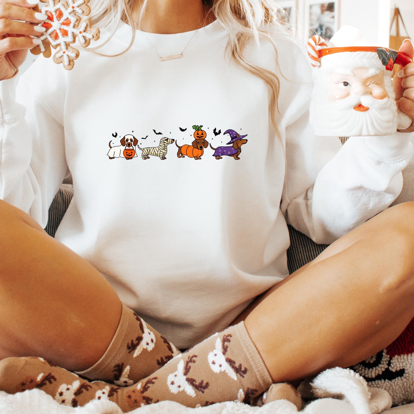 A person wearing a white sweater with a graphic of cartoon dogs and a Santa Claus mug, sitting on a couch with their legs crossed.