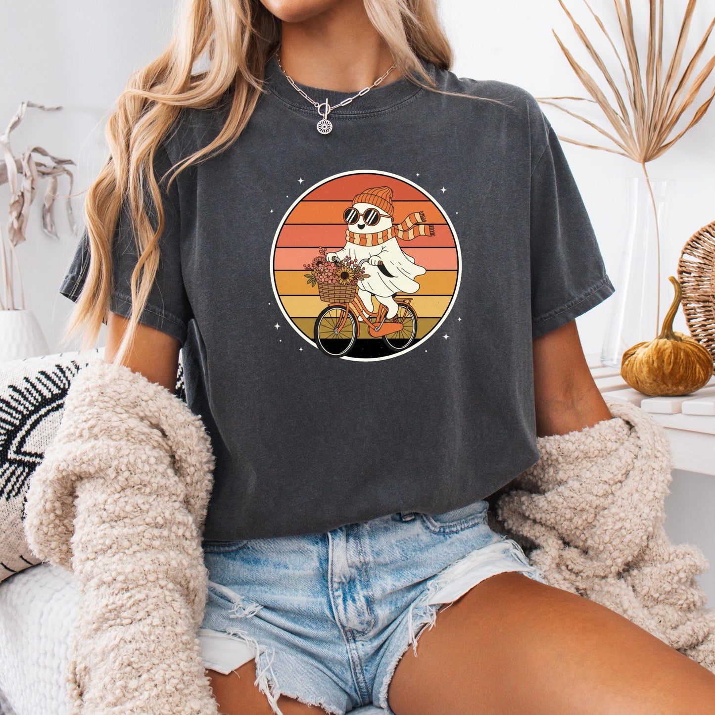 A woman wearing a black t-shirt with a graphic of a ghost riding a bicycle, along with a necklace and a beige sweater.