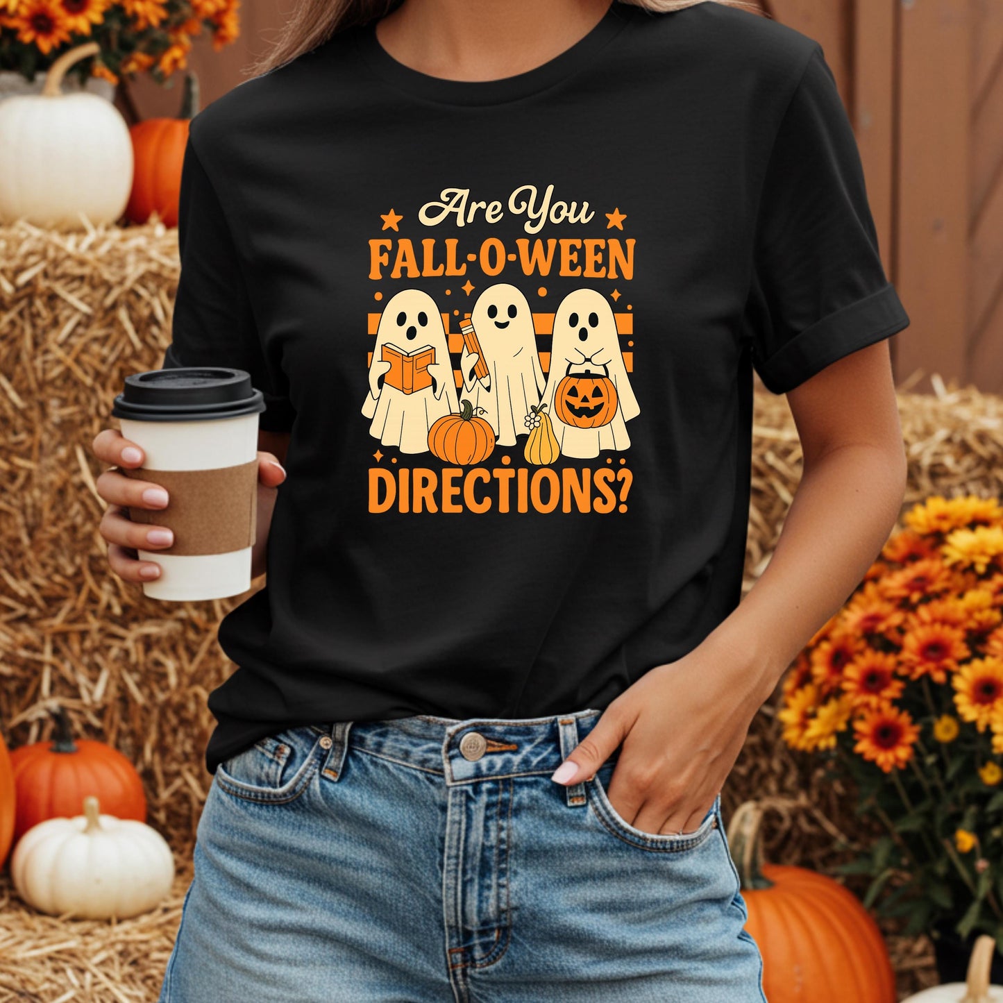 A person wearing a black t-shirt with a Halloween-themed graphic design, holding a coffee cup, and standing in front of a hay bale and pumpkins.
