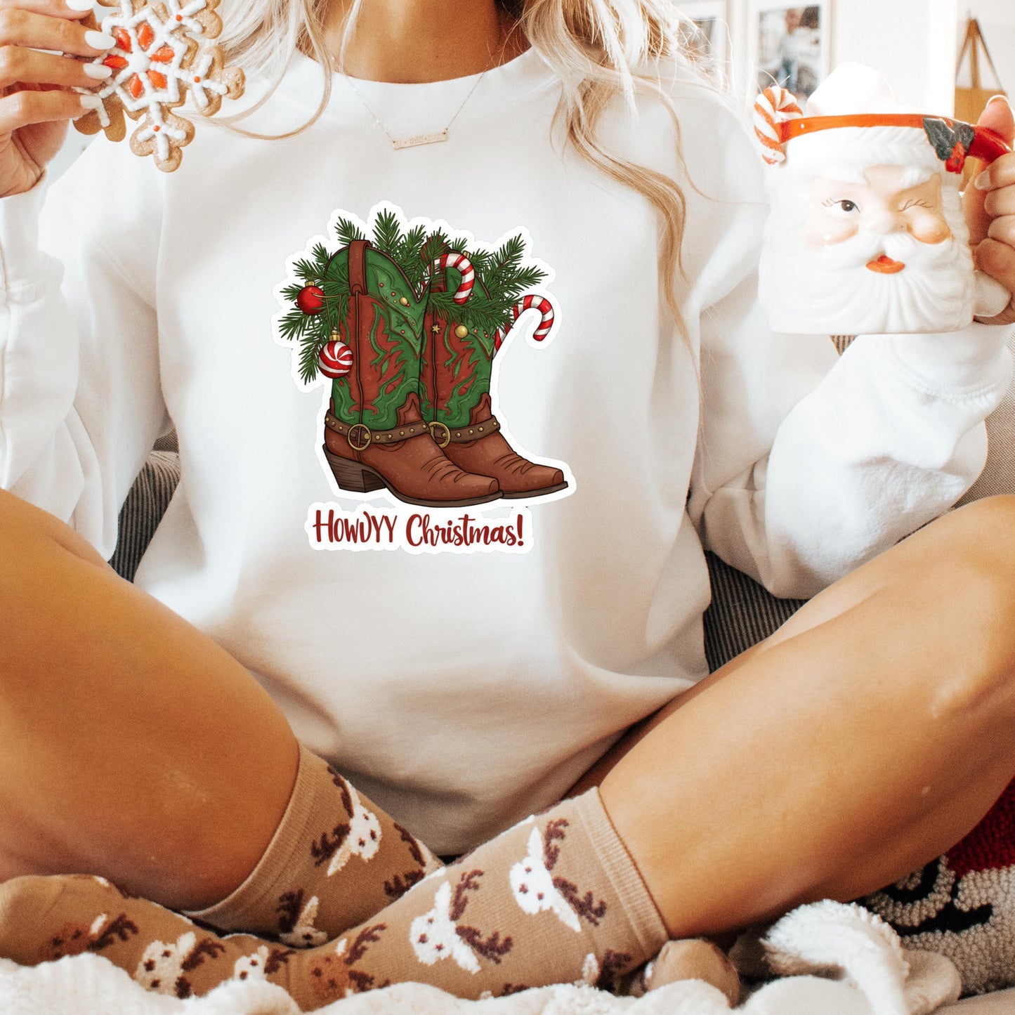 A person wearing a white sweater with a Christmas-themed graphic of cowboy boots and a Santa Claus mug, sitting on a couch with festive decorations around them.