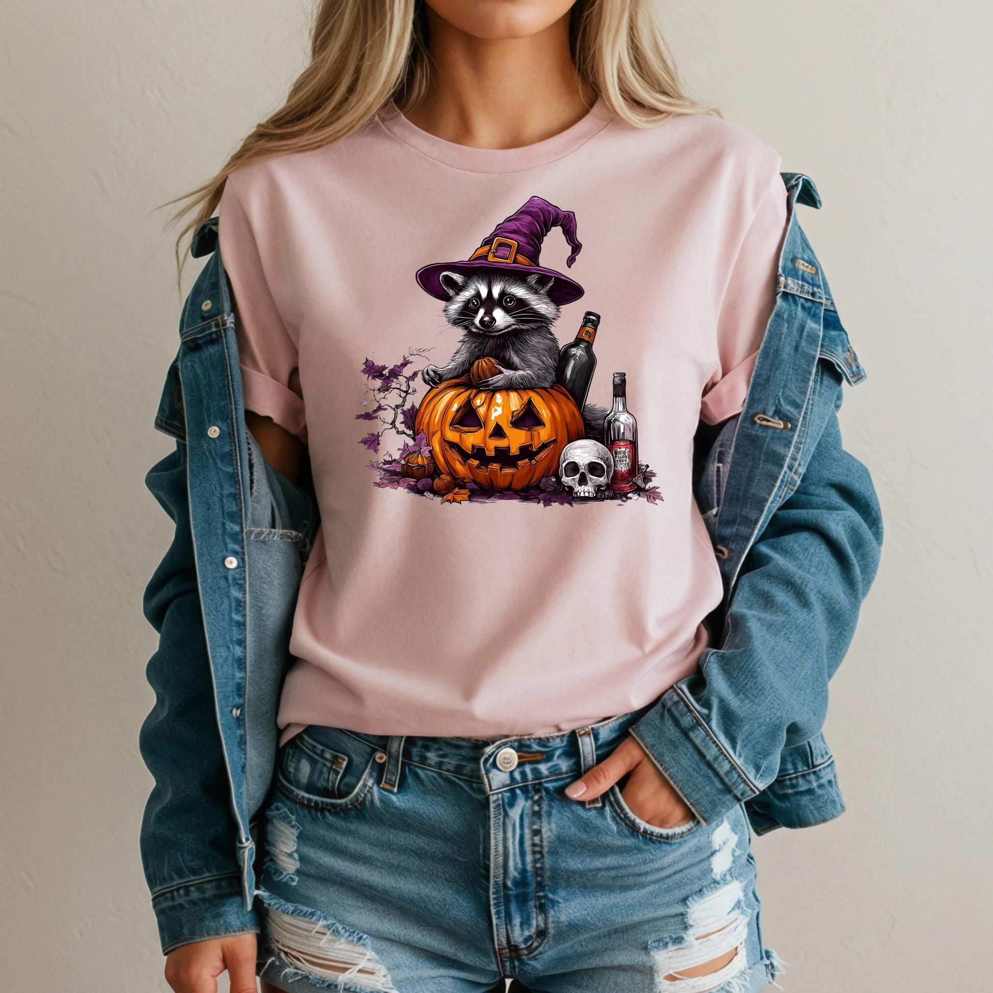 A woman wearing a pink t-shirt with a graphic of a raccoon wearing a witch's hat and holding a jack-o-lantern, along with other Halloween-themed elements.