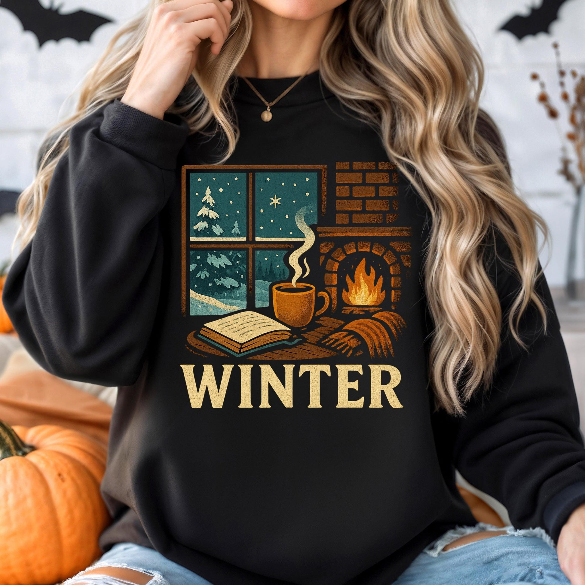 A woman wearing a black sweatshirt with a graphic design depicting a cozy winter scene, including a fireplace, a book, and a cup of coffee.