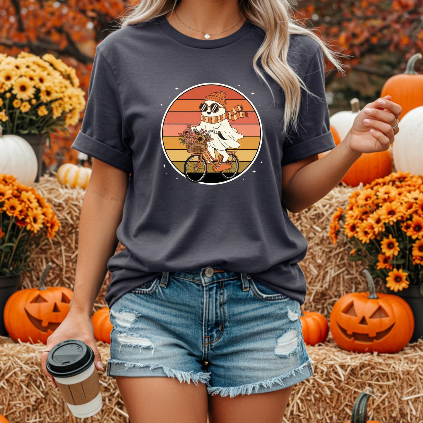 A woman wearing a t-shirt with a graphic of a ghost riding a bicycle is holding a coffee cup in front of a background of pumpkins and flowers.