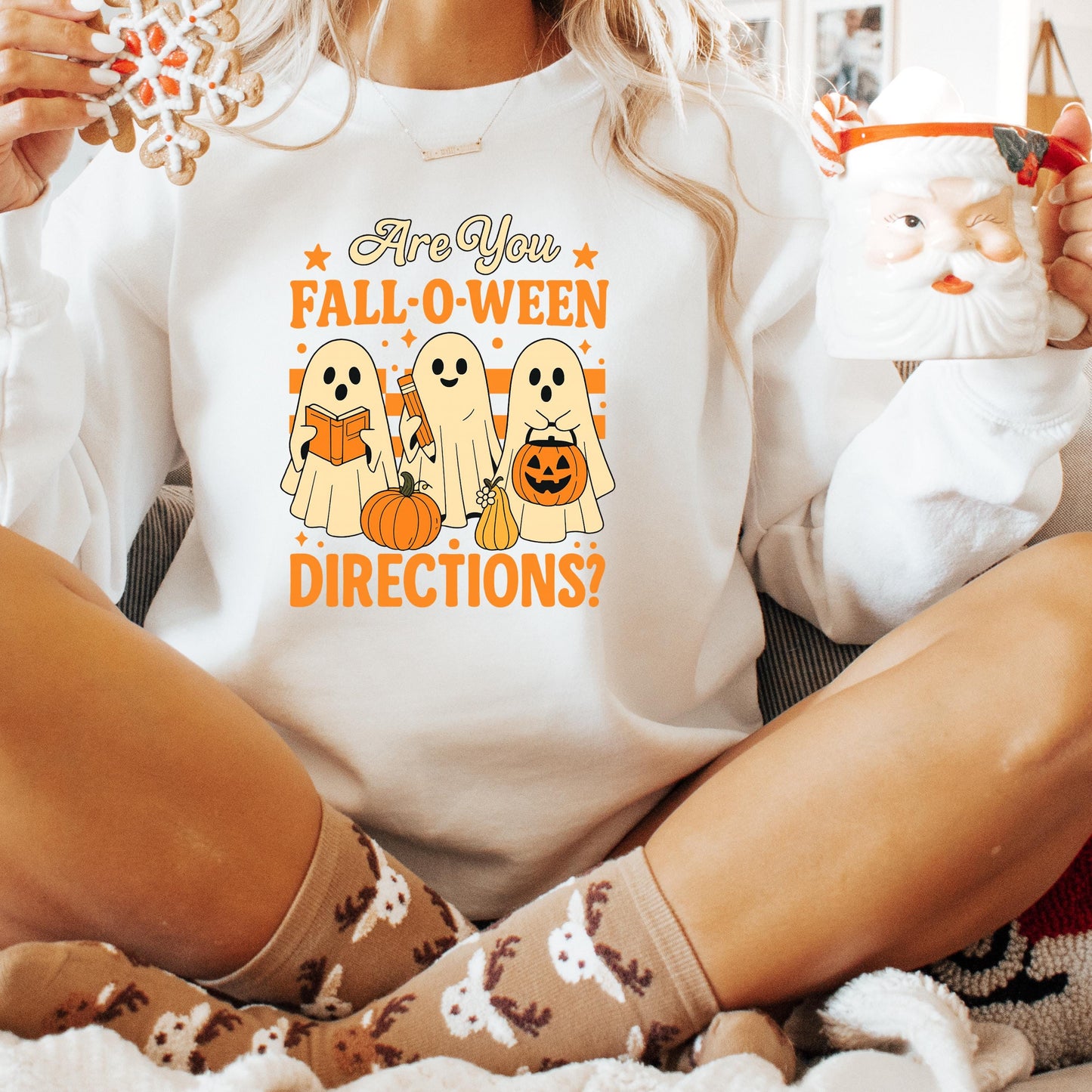 A person wearing a white sweatshirt with a Halloween-themed graphic and text, holding a mug and a small stuffed animal.