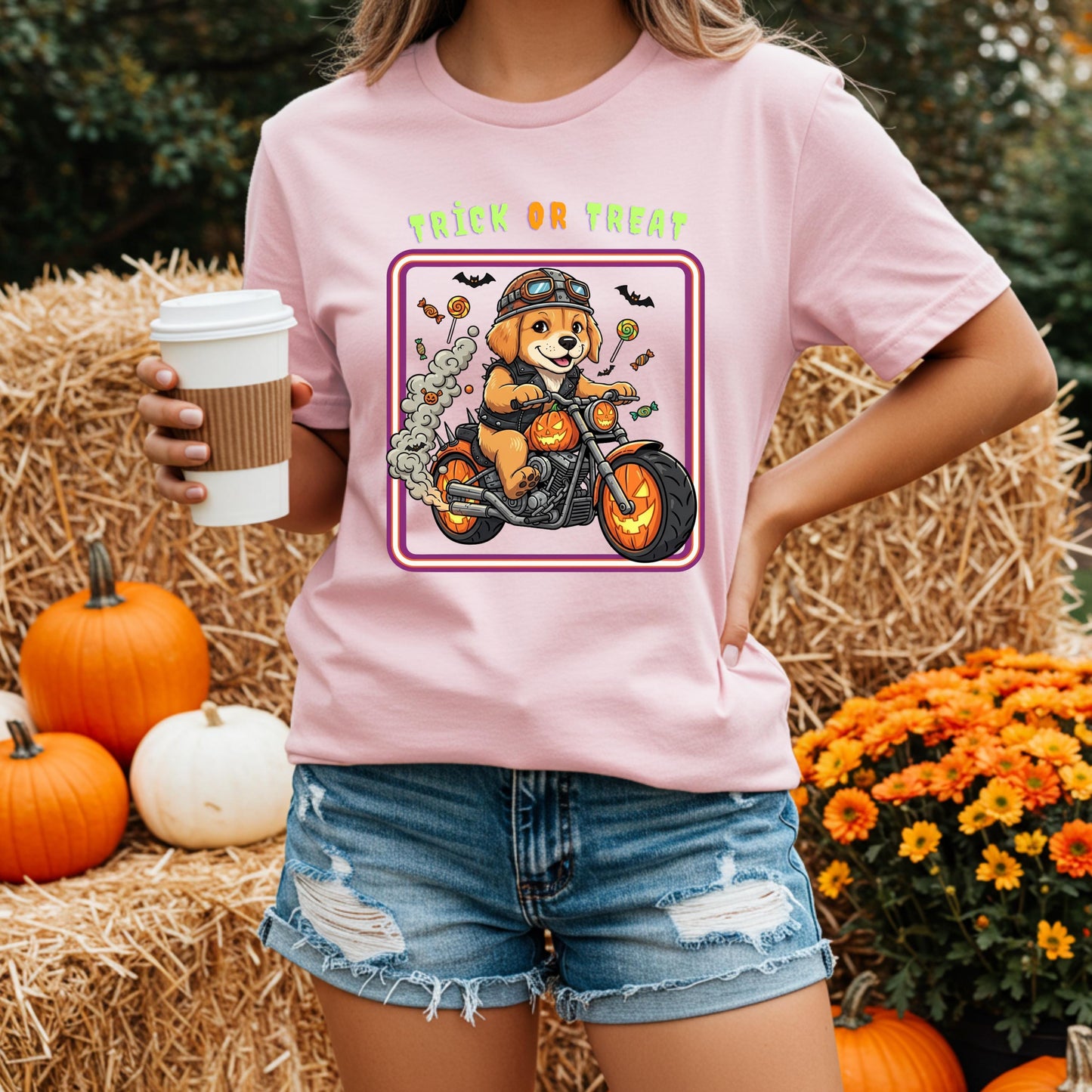 A woman wearing a pink t-shirt with a graphic of a dog riding a motorcycle and holding a coffee cup, standing in front of hay bales and pumpkins.