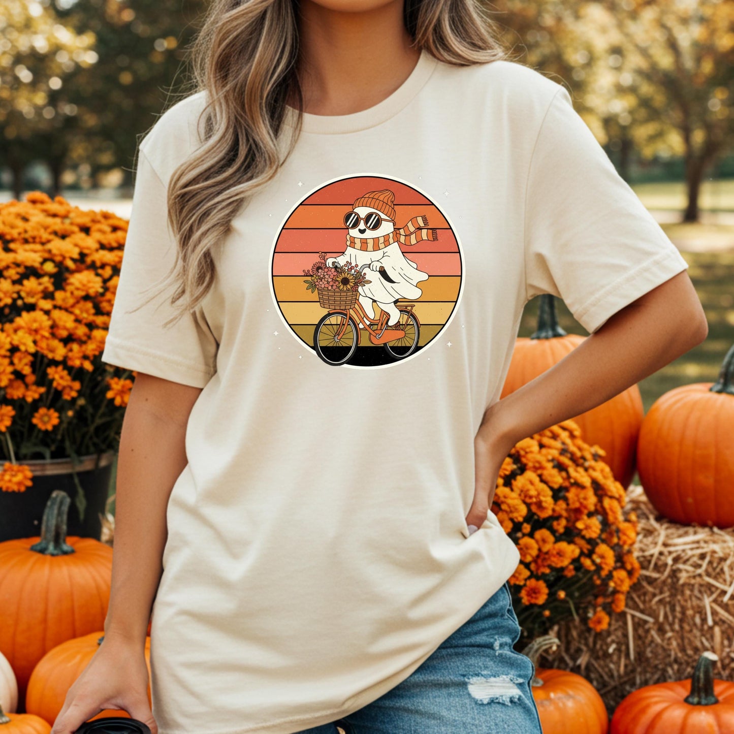 A woman wearing a white t-shirt with a graphic of a ghost riding a bicycle, surrounded by pumpkins and flowers in the background.