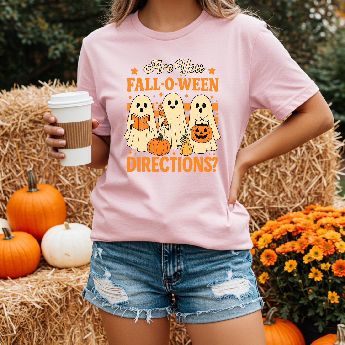 A woman wearing a pink t-shirt with a Halloween-themed graphic and holding a coffee cup, standing in front of a hay bale and pumpkins.