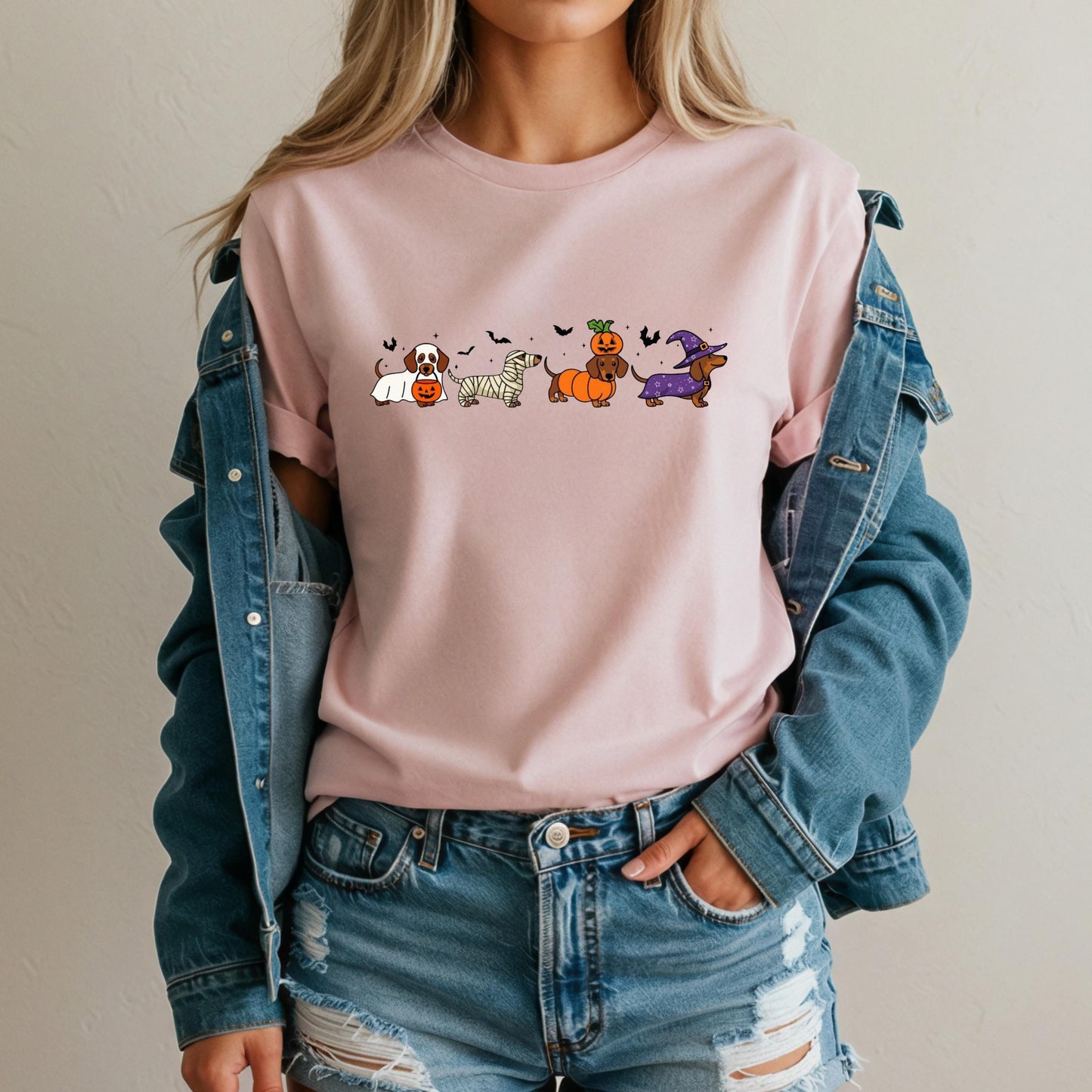 A woman wearing a pink t-shirt with a graphic of cartoon dogs and Halloween-themed accessories, paired with a denim jacket and distressed jeans.