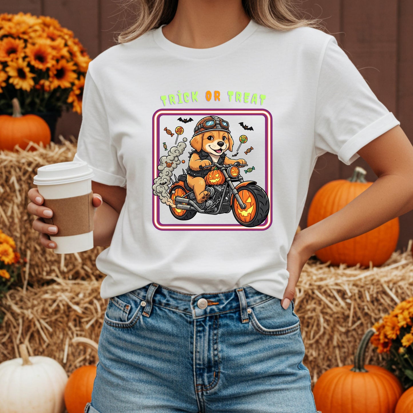 A woman wearing a white t-shirt with a graphic of a dog riding a motorcycle and the text &quot;Trick or Treat&quot; is holding a coffee cup in front of a hay bale and pumpkins.