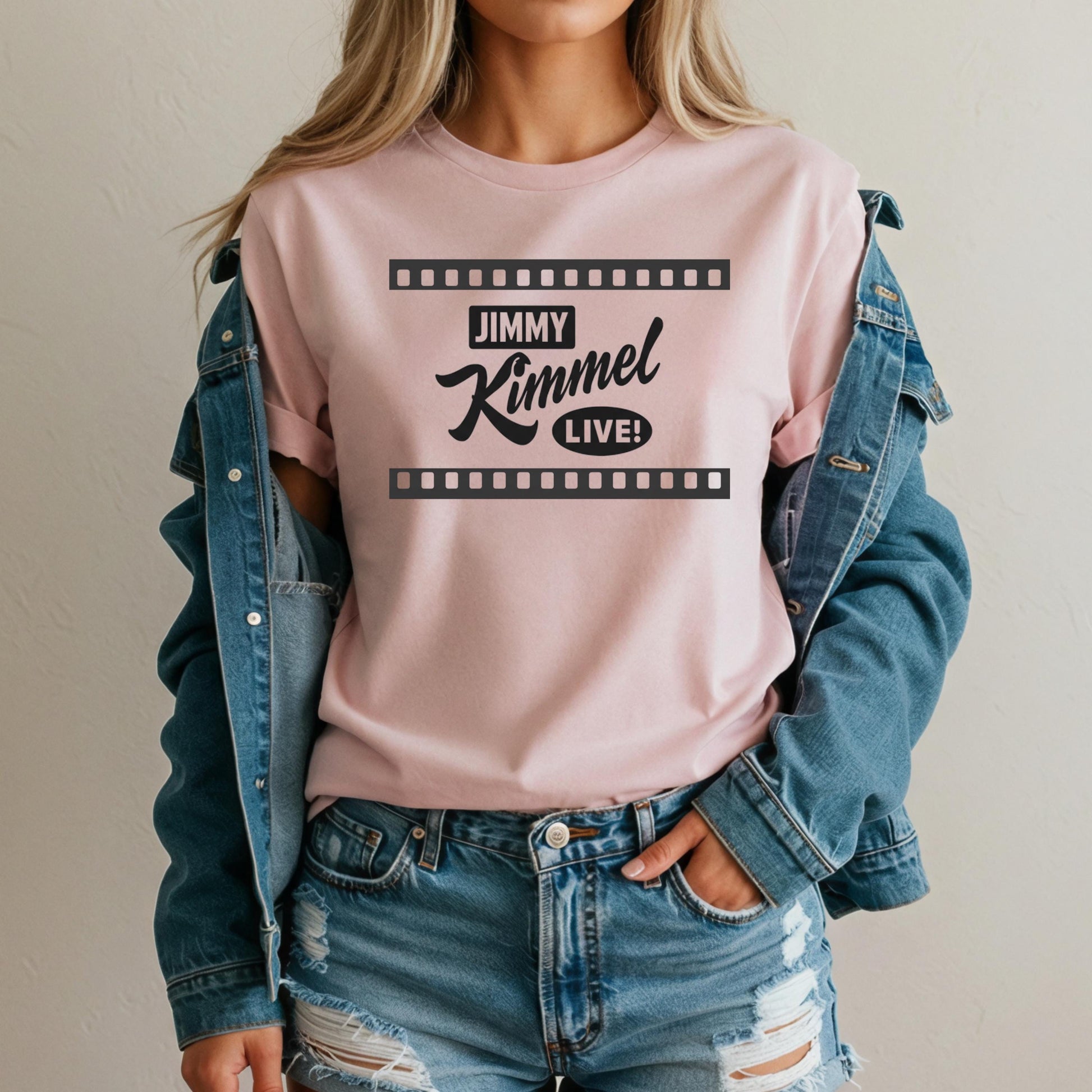 A woman wearing a pink t-shirt with the text &quot;JIMMY KIMMEL LIVE!&quot; printed on it, along with a denim jacket and distressed jeans.