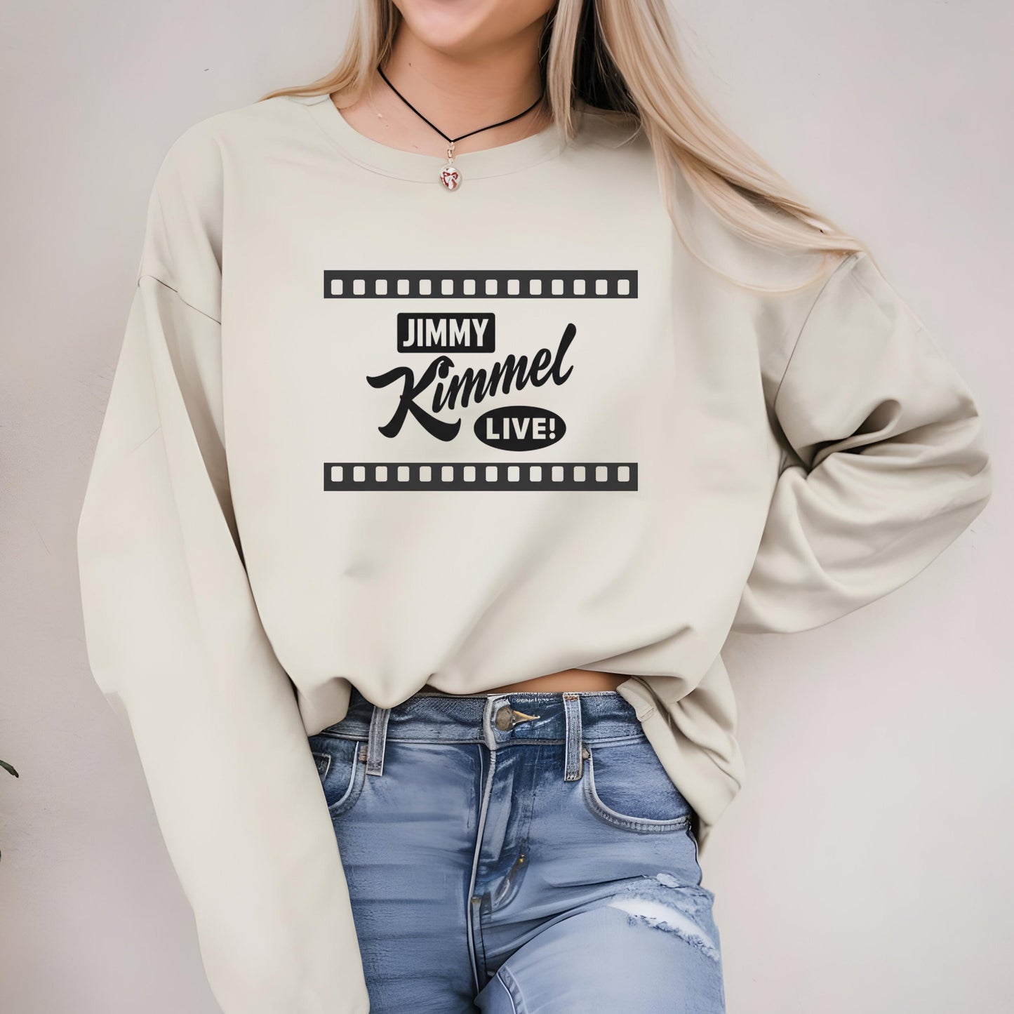 A woman wearing a white sweater with the text &quot;JIMMY KIMMEL LIVE!&quot; printed on it, along with a necklace. She is also wearing blue jeans.