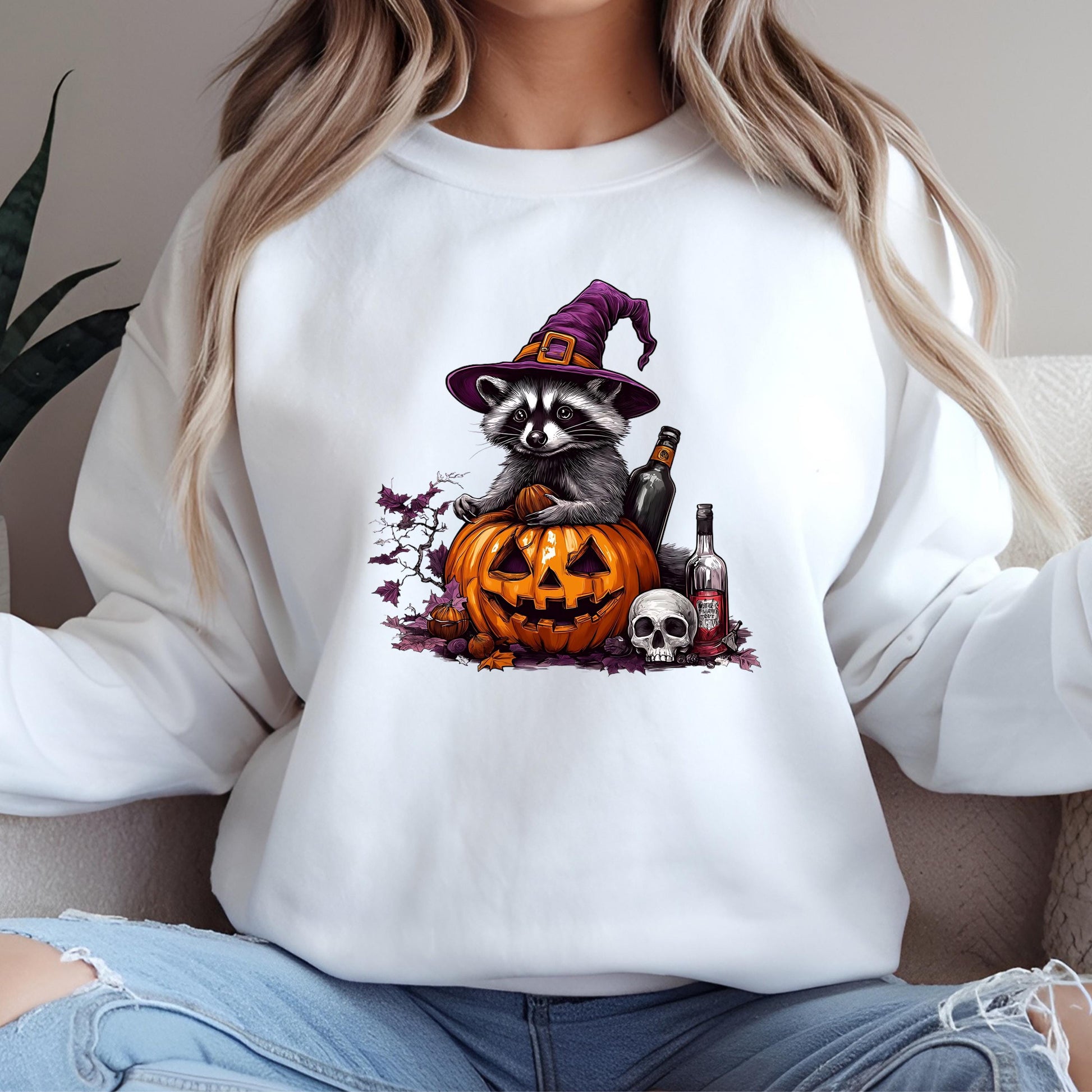 A white sweatshirt with a graphic design depicting a raccoon wearing a witch&#39;s hat, sitting on a carved pumpkin with a jack-o-lantern face, surrounded by Halloween-themed elements such as a skull, bottle, and bottle cap.
