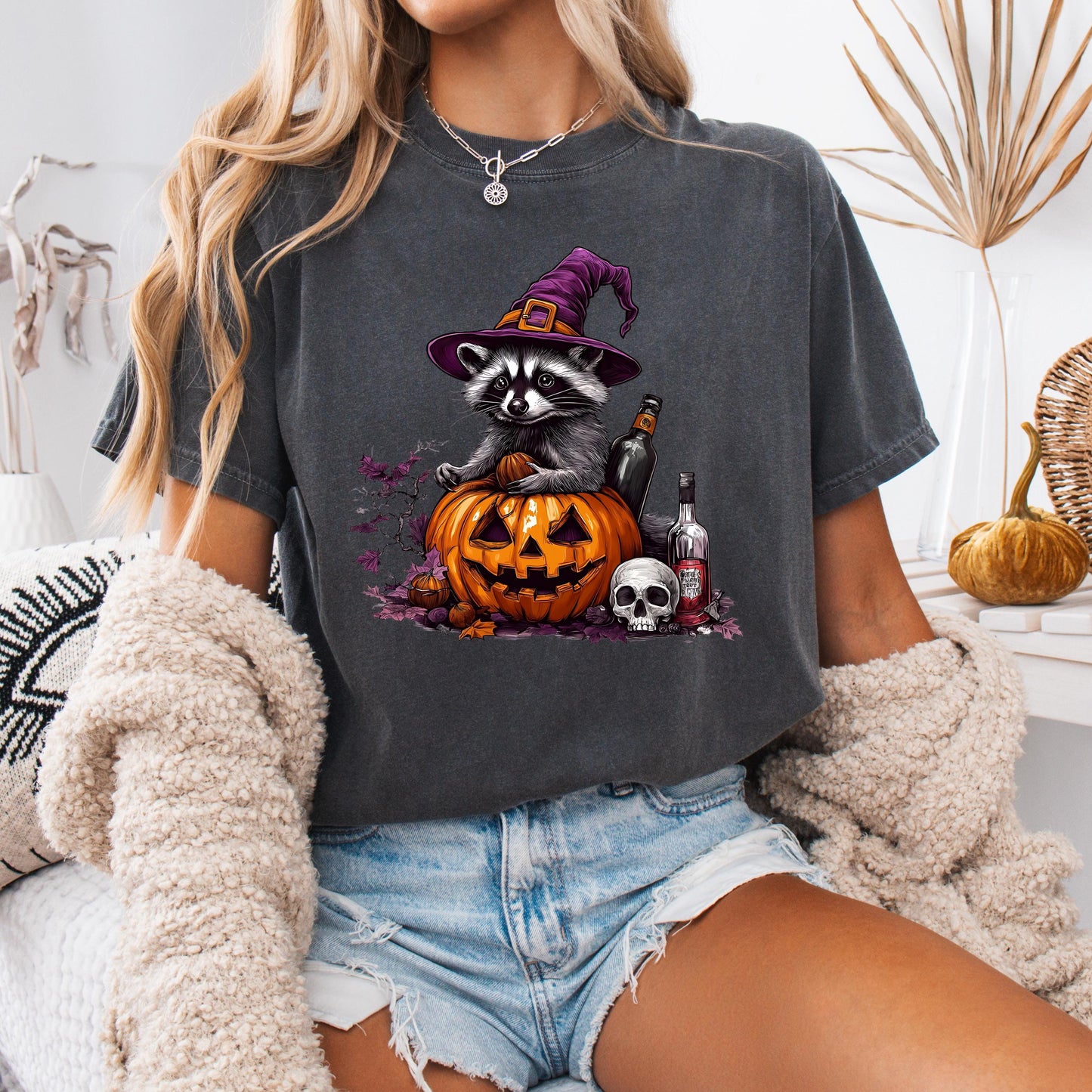 A woman wearing a gray t-shirt with a Halloween-themed graphic of a raccoon wearing a witch&#39;s hat and sitting on a carved pumpkin.