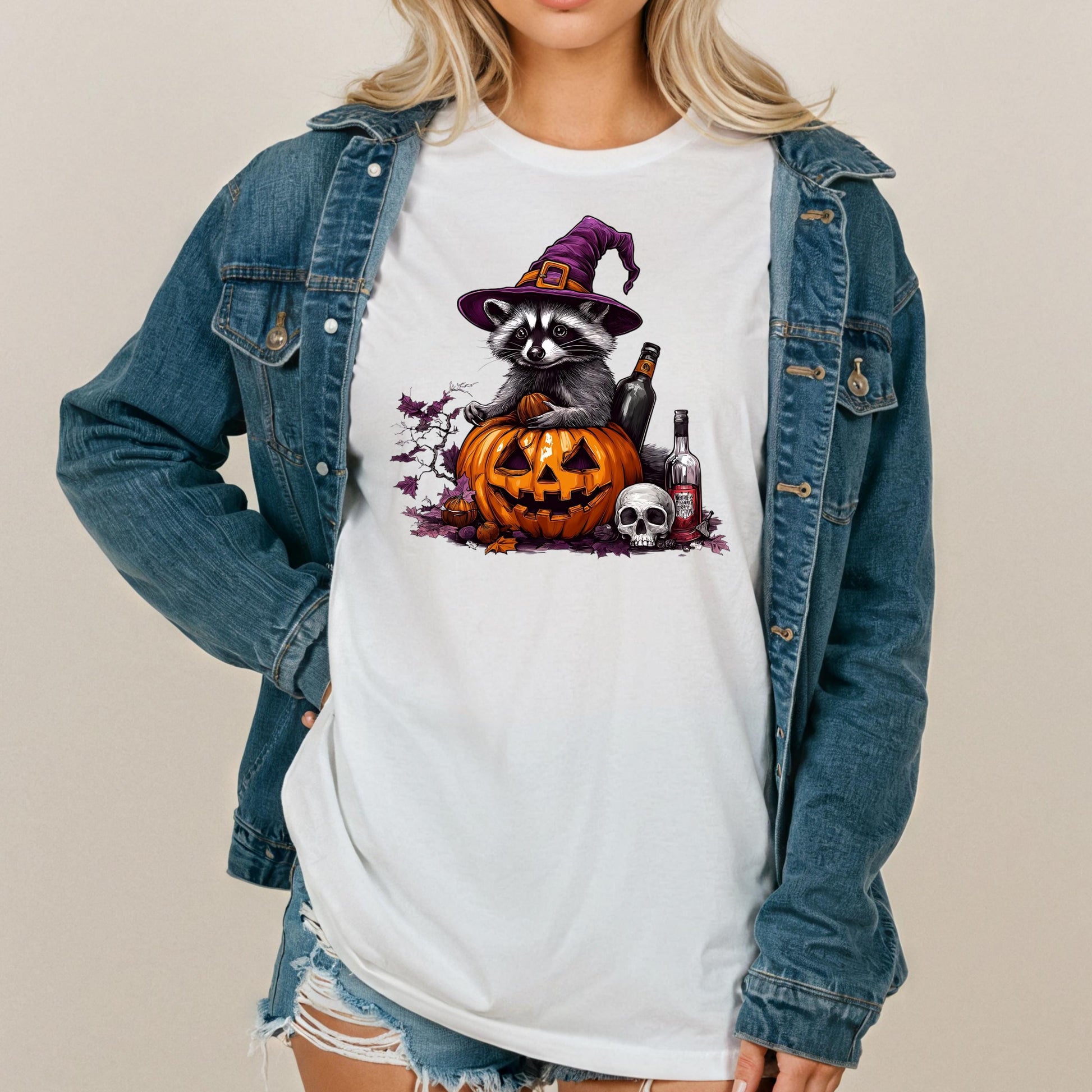 A woman wearing a denim jacket and a white t-shirt with a graphic design depicting a raccoon wearing a witch&#39;s hat and holding a jack-o-lantern, surrounded by Halloween-themed elements.