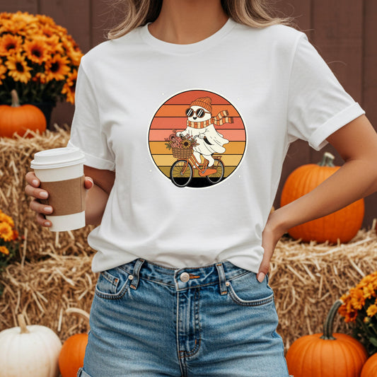 A woman wearing a white t-shirt with a graphic of a ghost riding a bicycle, holding a coffee cup, with orange pumpkins and a straw in the background.